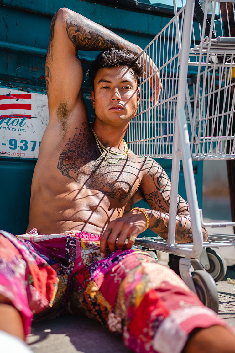 Topless man leans against a dumpster and shopping cart