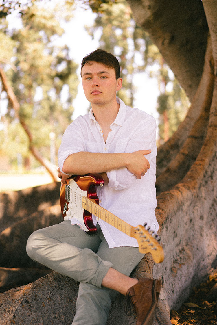 Man in white button-up shirt sits on a Moreton Bay fig tree with his electric guitar