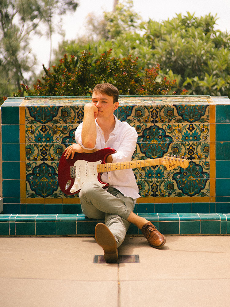 Man in white button-up shirt sits with electric guitar in front of a Spanish tile fountain